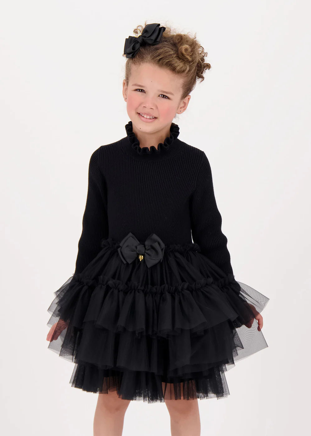 Elegant black ribbed jumper dress with layered soft tulle skirt and cotton lining. Features a frilled neck, signature bow, and gold heart charm on the waist.
