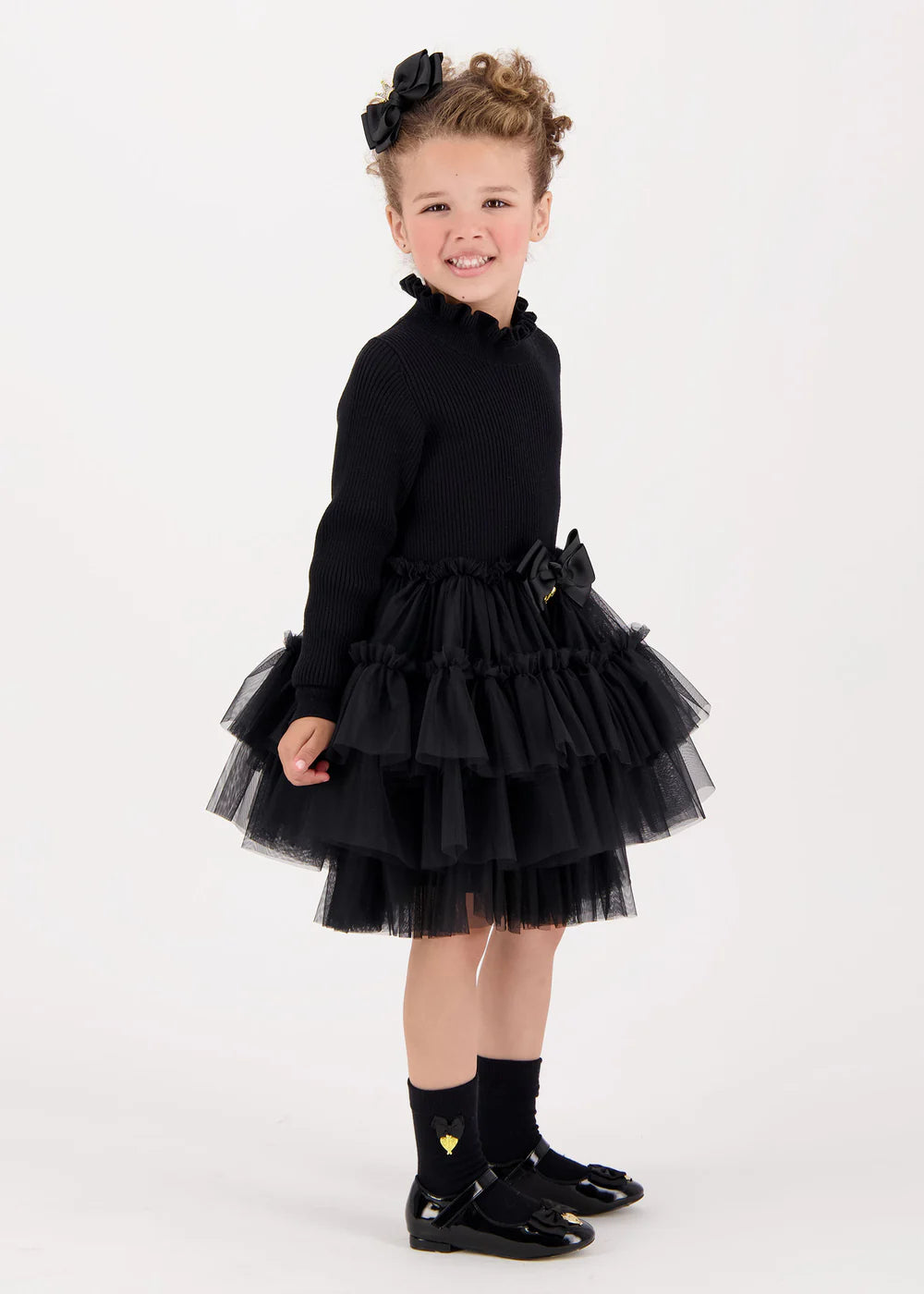 Elegant black ribbed jumper dress with layered soft tulle skirt and cotton lining. Features a frilled neck, signature bow, and gold heart charm on the waist.